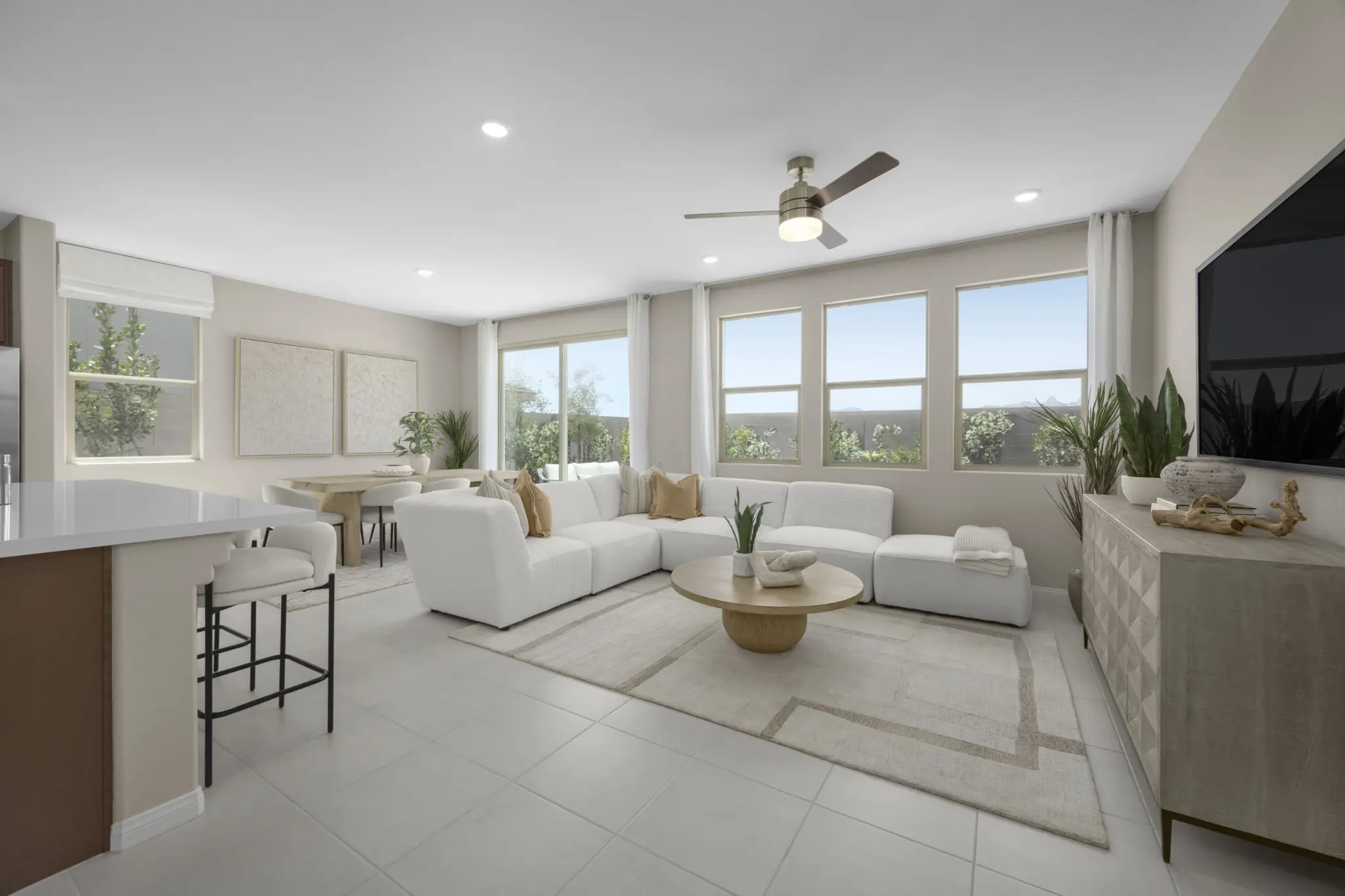 KBLV_LandingsatCaldwellPark_Plan1832-Greatroom_8321b-11×8-1-2048×1365 A modern, bright living room in Plan 1832 at Caldwell Park by KB Home features large windows, a white sectional sofa, round wooden coffee table, wall-mounted TV, dining area, and several potted plants on tiled floors.