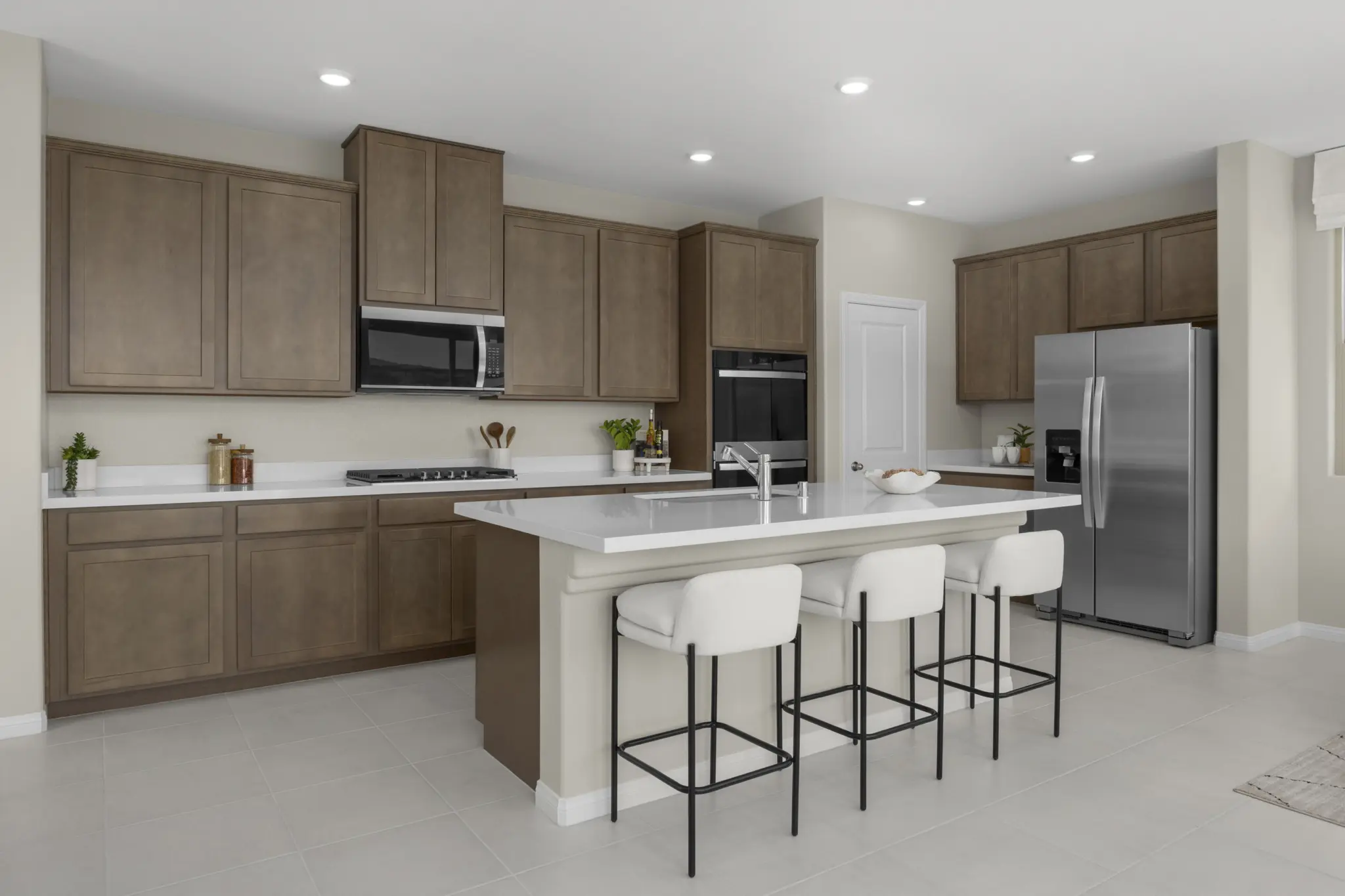KBLV_LandingsatCaldwellPark_Plan1832-Kitchen_7571c-11×8-1-2048×1365 Modern kitchen with light brown cabinets, stainless steel appliances, and a center island featuring a sink and three white cushioned bar stools. This bright space with recessed lighting is part of Plan 1832 at Caldwell Park by KB Home.