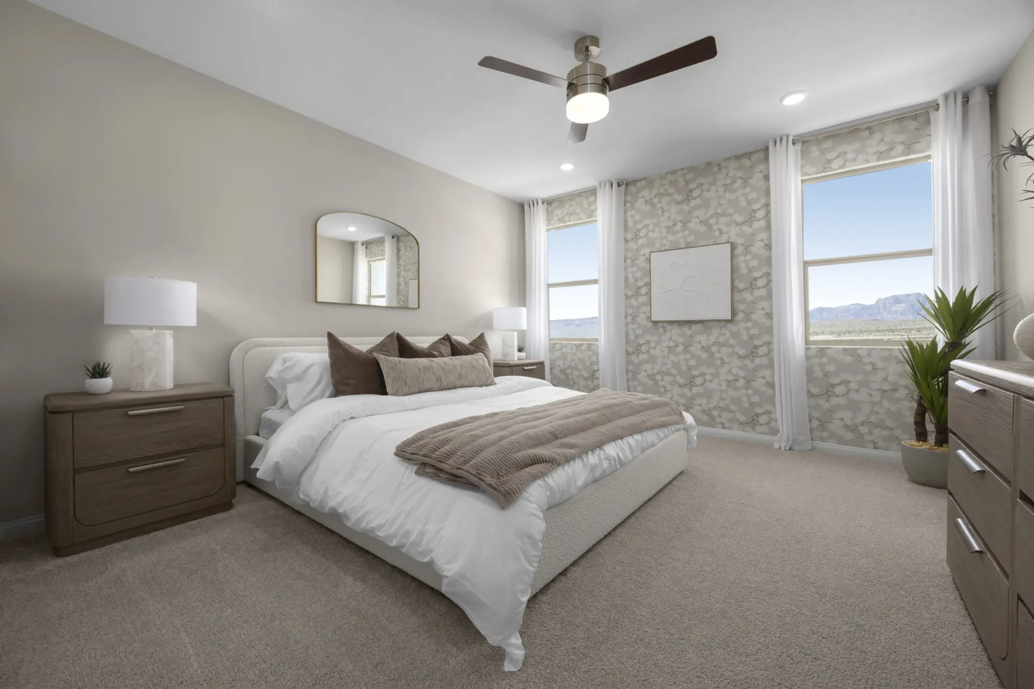 KBLV_LandingsatCaldwellPark_Plan1832-PrimaryBedroom_8454b-11×8-1-2048×1365 Modern bedroom in Plan 1832 at Caldwell Park by KB Home featuring a large bed with brown and white bedding, wooden nightstands and lamps, dresser, potted plant, ceiling fan, light carpet, and two windows with mountain views.
