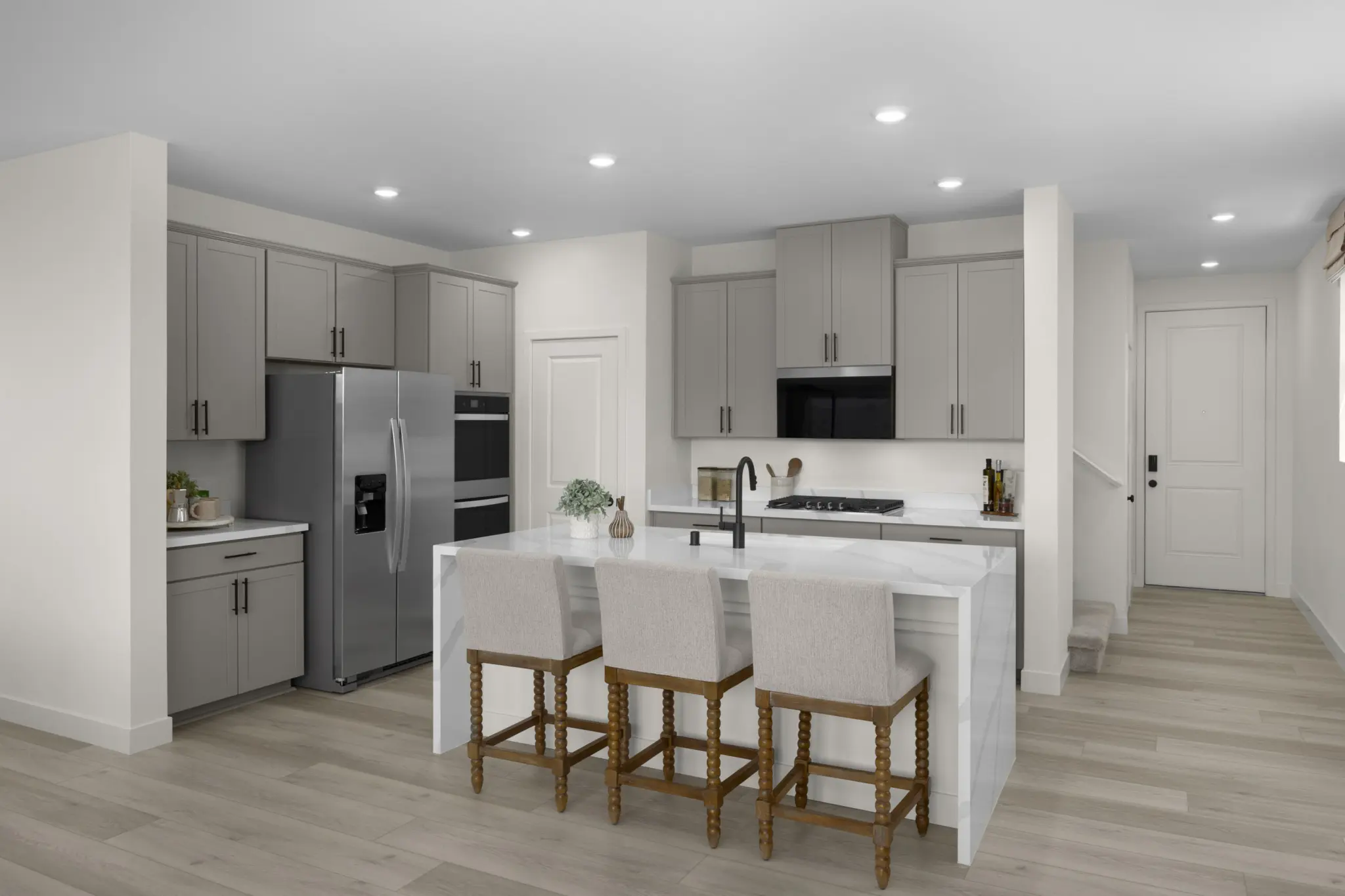 KBLV_LandingsatCaldwellPark_Plan2070-Kitchen_7375b-11×8-1-2048×1365 Modern kitchen in Plan 2070 at Caldwell Park by KB Home features gray cabinets, stainless appliances, a marble island with sink, and three upholstered bar stools. Recessed lighting and light wood floors create a bright, clean atmosphere.