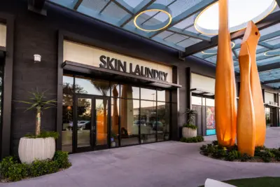 Storefront of Skin Laundry with large glass doors and windows, modern black and beige exterior, and tall orange abstract sculptures in front on a landscaped sidewalk.