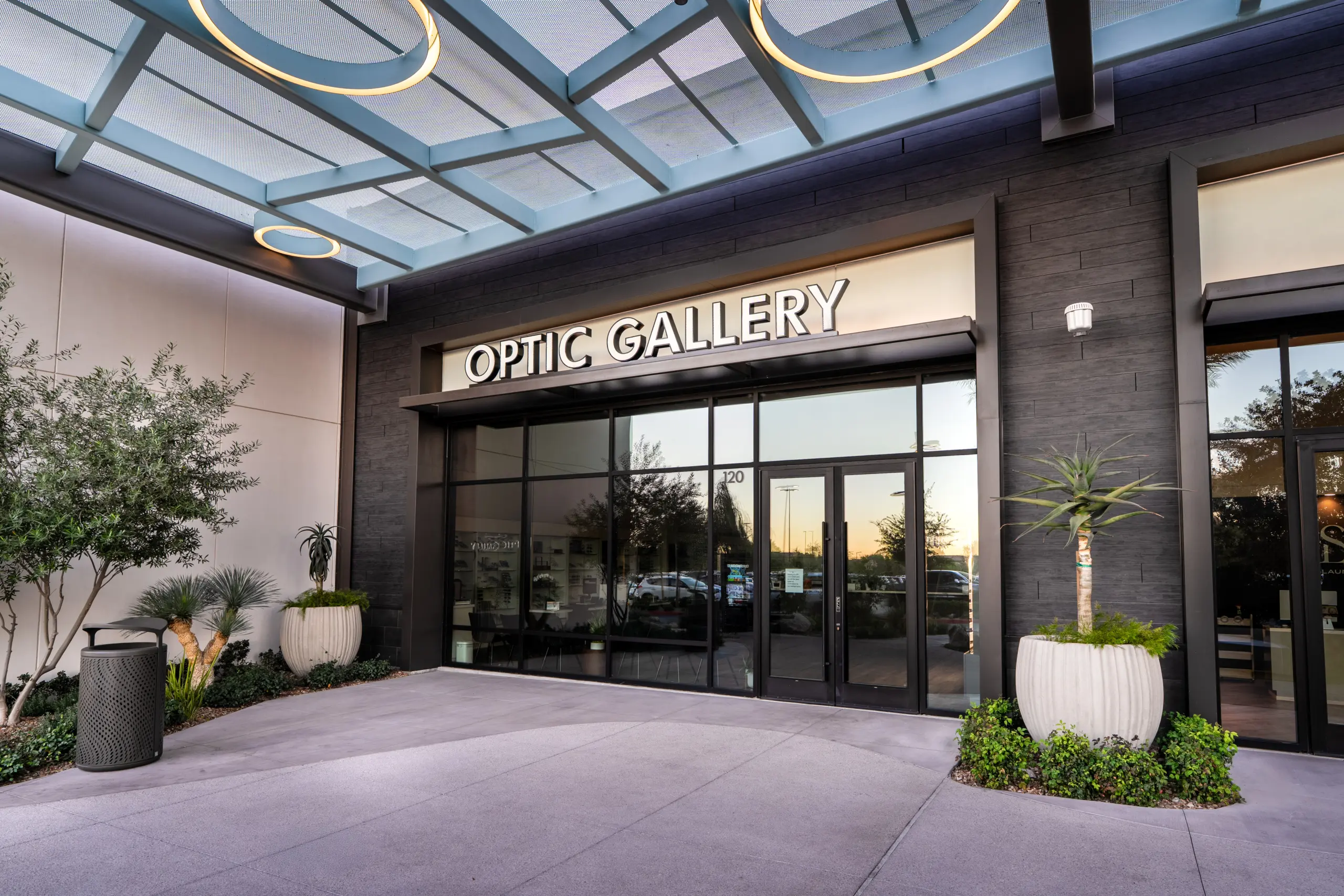 DSC05986 Entrance to a modern building with large glass doors and a sign reading Optic Gallery above. There are potted plants on either side of the doorway and a metal trash can to the left.