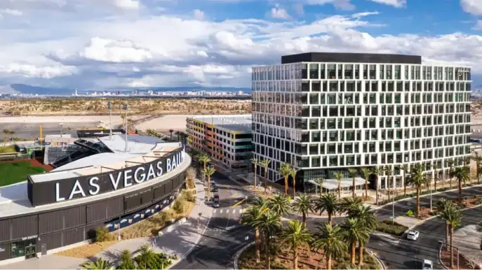 1700Pavilion A modern city scene with the Las Vegas Ballpark in the foreground, a tall glass office building nearby, palm trees, roads, and the Las Vegas skyline visible in the distance under a partly cloudy sky.