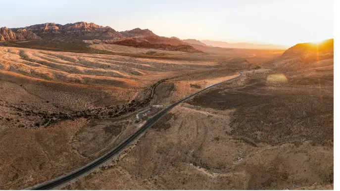 explore-featured Aerial view of a long, winding road cutting through a vast, arid desert landscape with rocky hills in the distance and the sun setting on the horizon, casting a warm golden light.