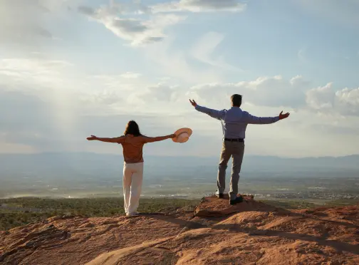 Custom_Homesites A man and woman stand on a rocky hilltop with arms outstretched, facing a scenic view of open landscape and distant mountains under a partly cloudy sky.