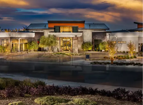 A modern, single-story building with large windows and a landscaped entrance is illuminated at dusk, with dramatic clouds in the sky and a driveway in front.