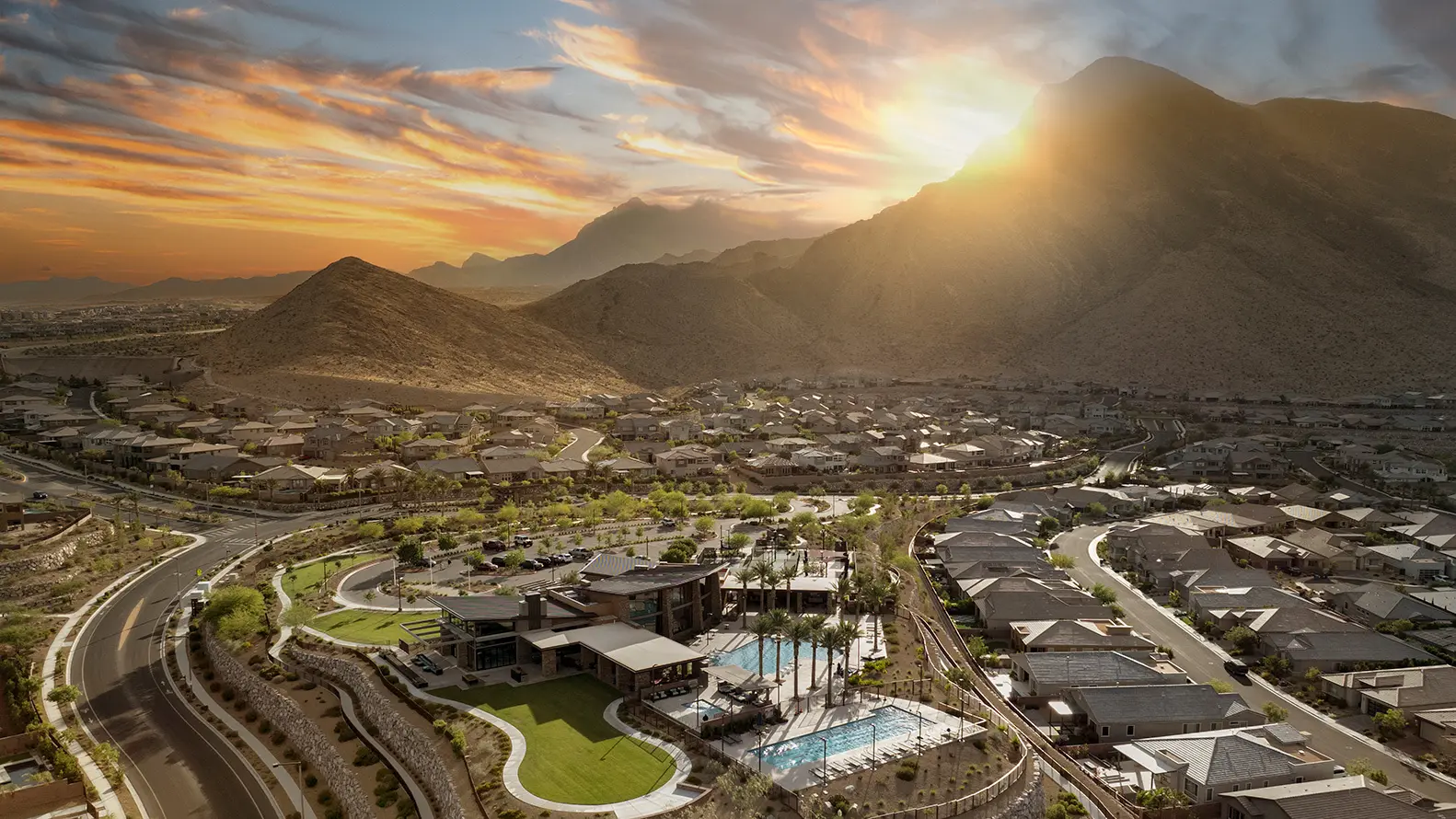 summerlin-about-mm A suburban neighborhood nestles beside desert mountains at sunset, with houses, winding roads, green parks, and a swimming pool, all illuminated by the golden sunlight behind the peaks.