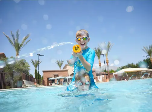summerlin-home_AMENITIES Two children playing in a swimming pool, with one child squirting water from a toy and both wearing swim goggles under a clear blue sky, palm trees and lounge chairs visible in the background.