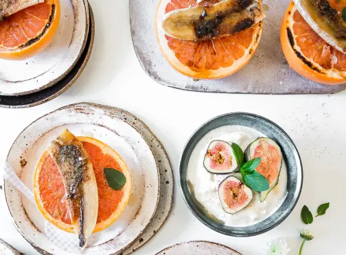 summerlin-home_DINING Several plates with broiled grapefruit halves topped with caramelized banana slices, and a bowl of yogurt garnished with fresh fig slices and basil, arranged on a white table.