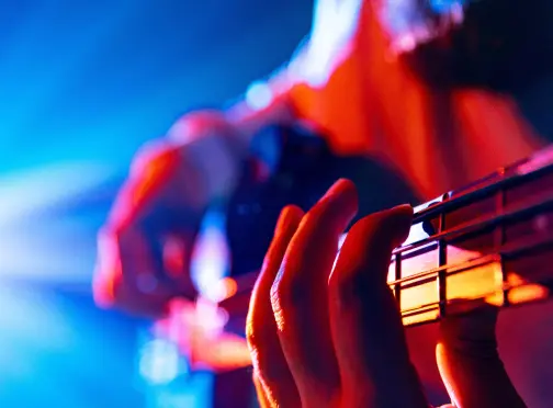 summerlin-home_EVENTS Close-up of a person playing an electric guitar, with fingers pressing the strings on the fretboard. The background features vibrant blue and orange stage lighting.