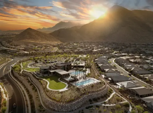 summerlin-home_HOMES Aerial view of a residential neighborhood at sunset, surrounded by mountains. The community features a clubhouse, green lawns, and a swimming pool with lounge chairs. The sun is setting behind the hills, casting a warm glow.