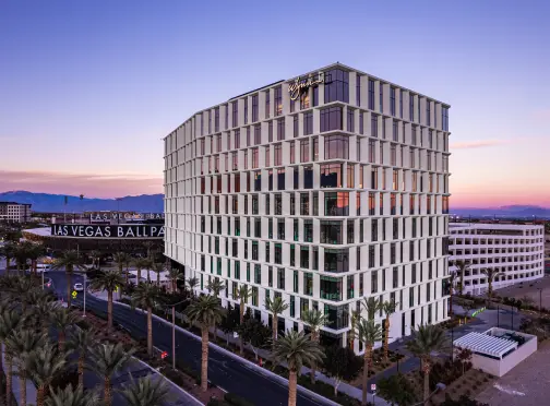 summerlin-home_OFFICES’ A modern, multi-story hotel building with a grid-like facade stands at sunset in Las Vegas, with palm trees lining the street and mountains visible in the background. A sign reads Las Vegas Ballpark.