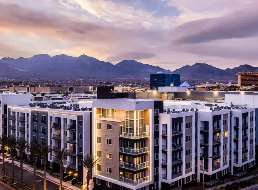 summerlin-home_RESIDENCES Modern mid-rise apartment buildings with lit balconies at dusk, palm trees in front, and mountains in the background under a colorful, cloudy sky.