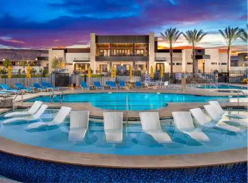 A modern resort pool area at sunset features in-pool lounge chairs, circular pools, palm trees, and a contemporary building in the background, with vibrant blue water and a colorful sky.