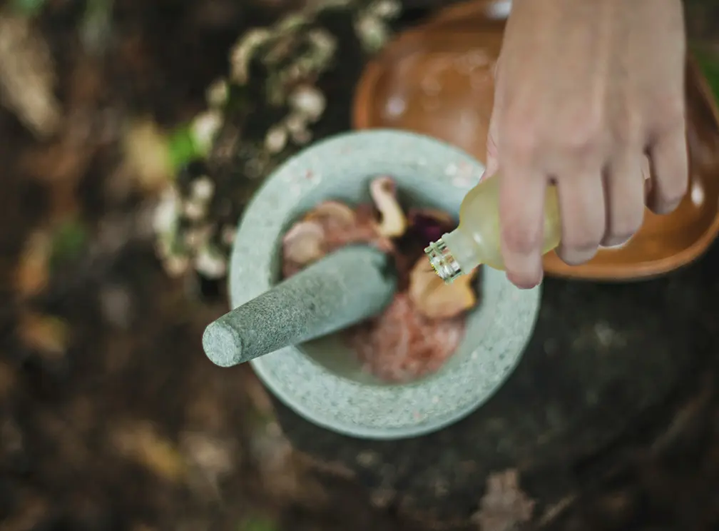 A hand pours liquid from a small bottle into a stone mortar containing dried herbs and roots, with a pestle resting inside, set outdoors on a natural surface.