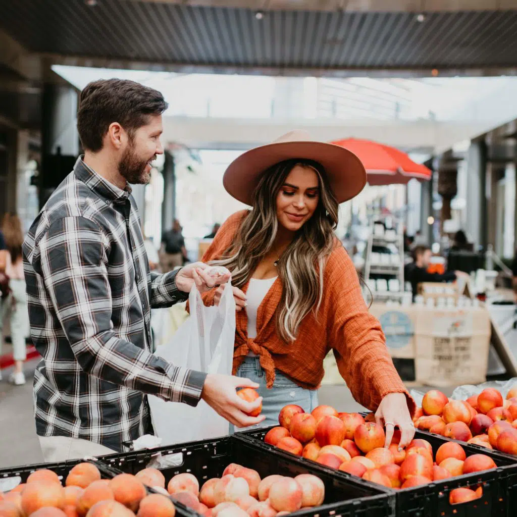 A man and woman select apples from outdoor market crates. The woman wears a wide-brim hat and an orange top, while the man holds a bag and wears a plaid shirt. Other shoppers and stands are visible in the background.