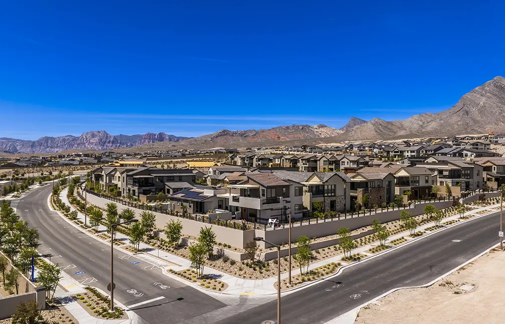A suburban neighborhood with modern houses, tree-lined streets, and minimal traffic, set against a backdrop of mountains under a clear blue sky. Desert landscaping surrounds the area.