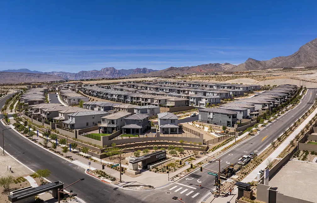 Aerial view of a modern suburban neighborhood with rows of similar gray houses, landscaped yards, and winding streets, set against a backdrop of mountains under a clear blue sky.