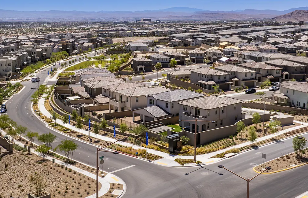 Aerial view of a suburban neighborhood with rows of modern houses, landscaped yards, and curving streets. The area appears dry and is surrounded by mountains in the distance under a clear blue sky.