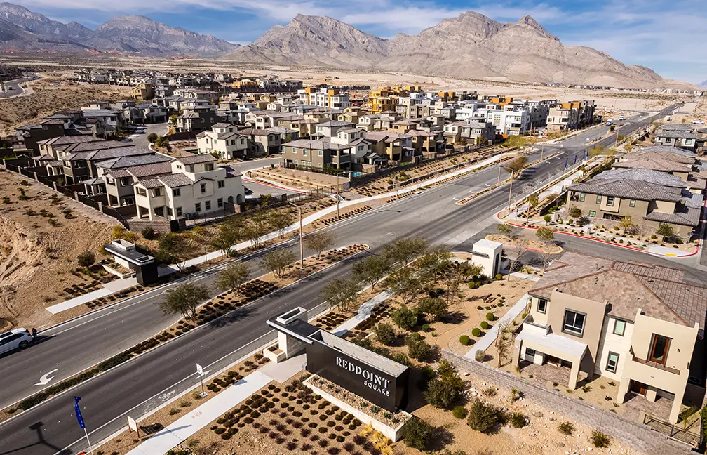 Aerial view of a suburban neighborhood with rows of modern houses, landscaped yards, and wide streets. A sign reading REDPOINT marks the entrance, with desert mountains and dry terrain in the background.