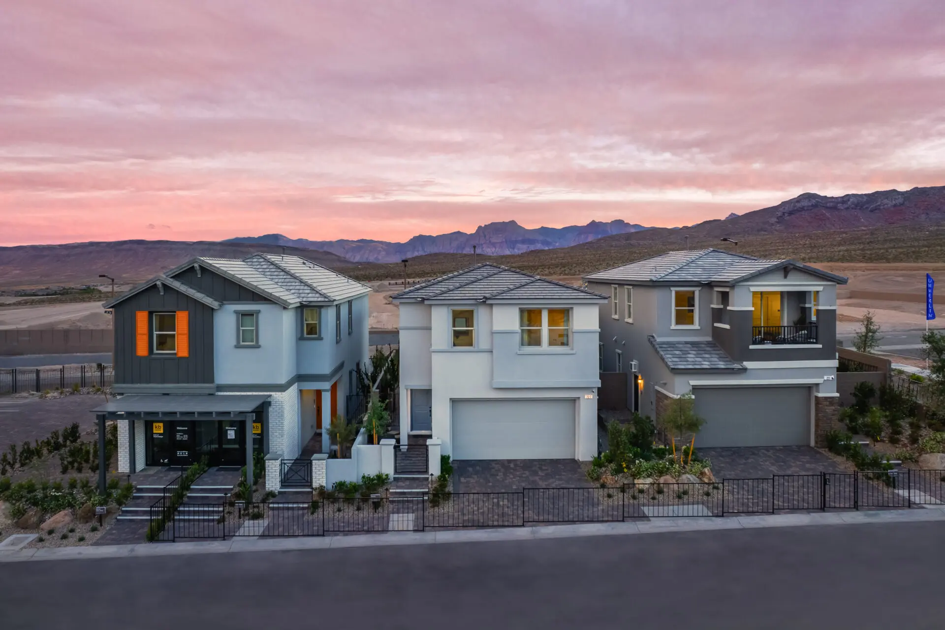 Summerlin Caldwell Park Three modern two-story houses with garages and landscaped yards are lined up on a street at sunset, with mountains and a colorful sky visible in the background.