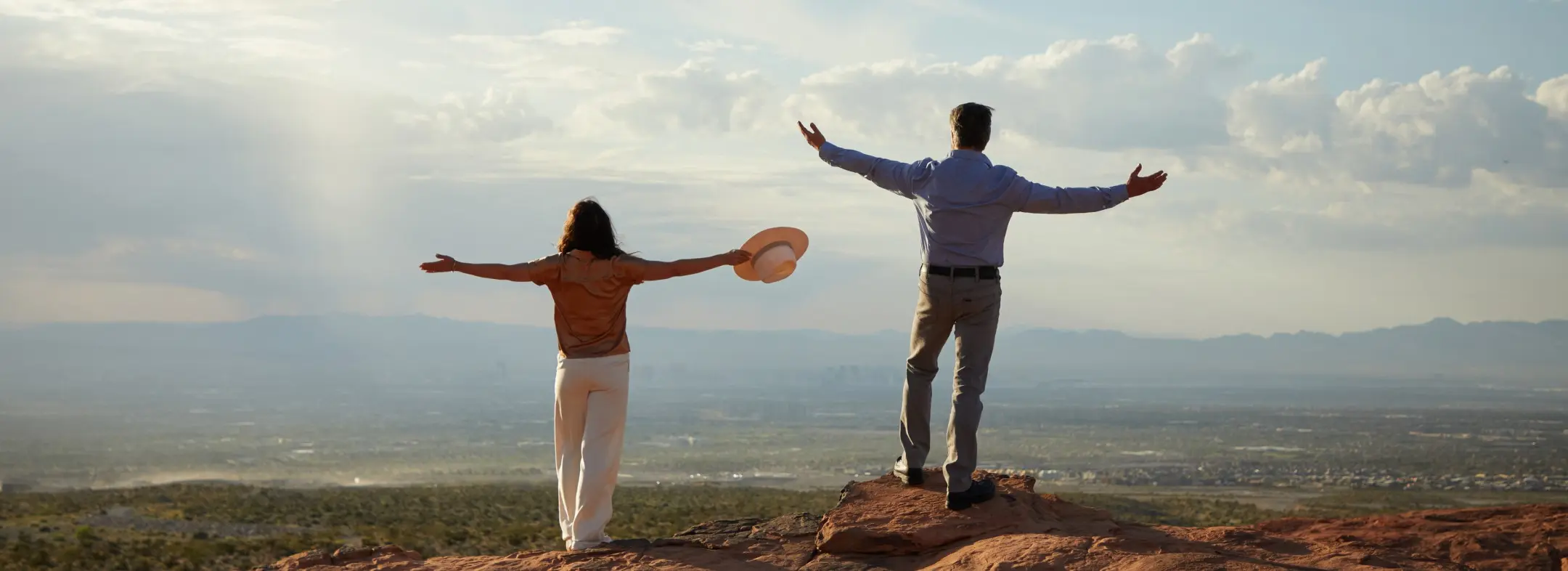 Two people stand on a rocky hilltop with arms outstretched, looking out over a vast landscape under a partly cloudy sky. One holds a hat, and both appear to be enjoying the expansive view.