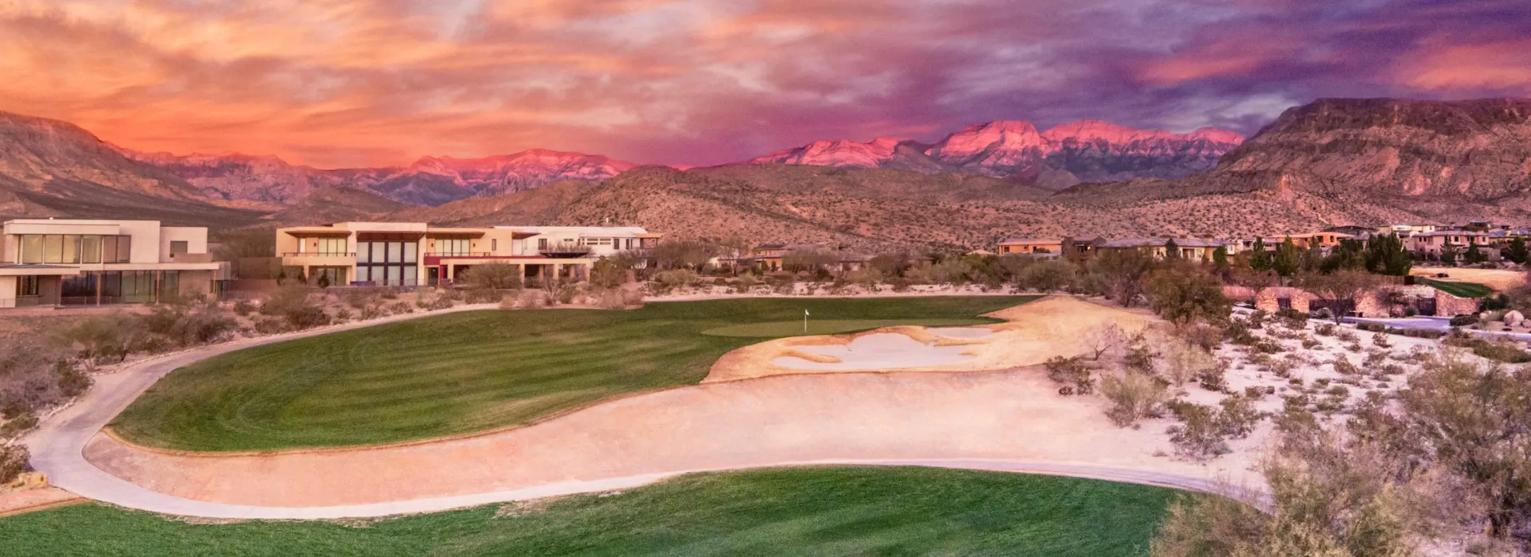 A golf course with sand bunkers and green grass is set against desert terrain and modern buildings. Purple and orange sunset clouds color the sky, and distant mountains glow pink in the background.