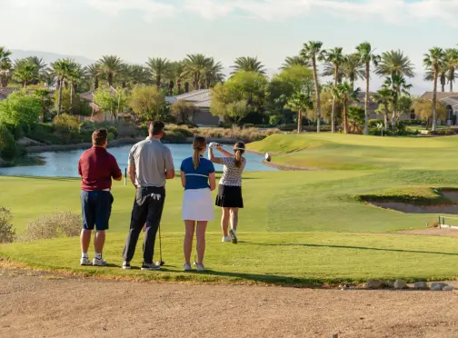 Four people stand on a golf course tee box, watching as one person swings a club. Palm trees and houses are visible in the background under a clear sky.