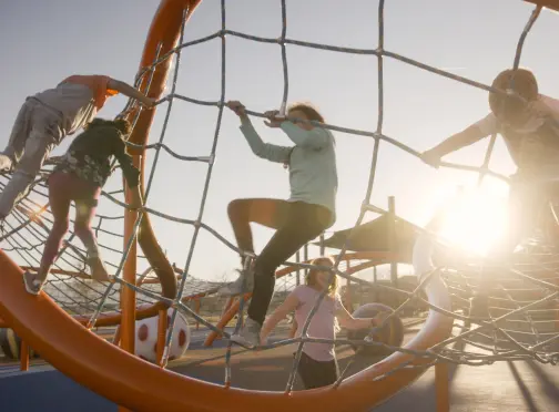 Summerlin_Explore-Parks Children climb and play on a large orange rope climbing structure at a playground, with the sun shining brightly in the background.