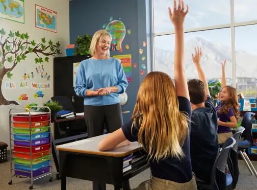 A smiling teacher stands at the front of a colorful classroom as several students sitting at desks raise their hands to answer a question. A large window shows a mountain view outside.