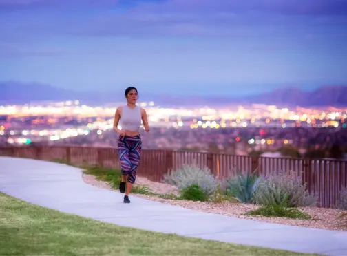 Summerlin_Explore-Trails A woman runs on a paved path in a park at dusk, with city lights glowing in the blurred background and mountains in the distance. She wears a white tank top and colorful leggings.