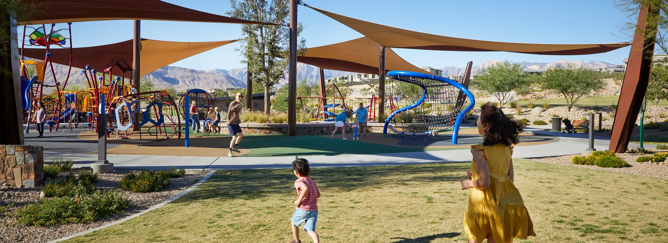 Summerlin_Parks Children and adults enjoy a sunny day at a modern playground with colorful climbing structures and shade sails, set in a park with mountains visible in the background. A woman and child walk towards the play area.