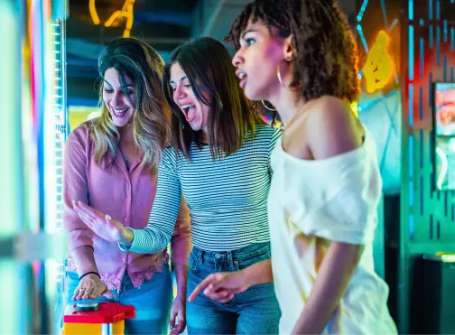 Three young women excitedly play an arcade game together, with one pressing a large button while the others cheer. Bright, colorful lights and arcade machines fill the background.