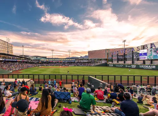 Fans sit on a grassy hill watching a baseball game in a modern stadium at sunset, with city buildings and a colorful sky in the background.
